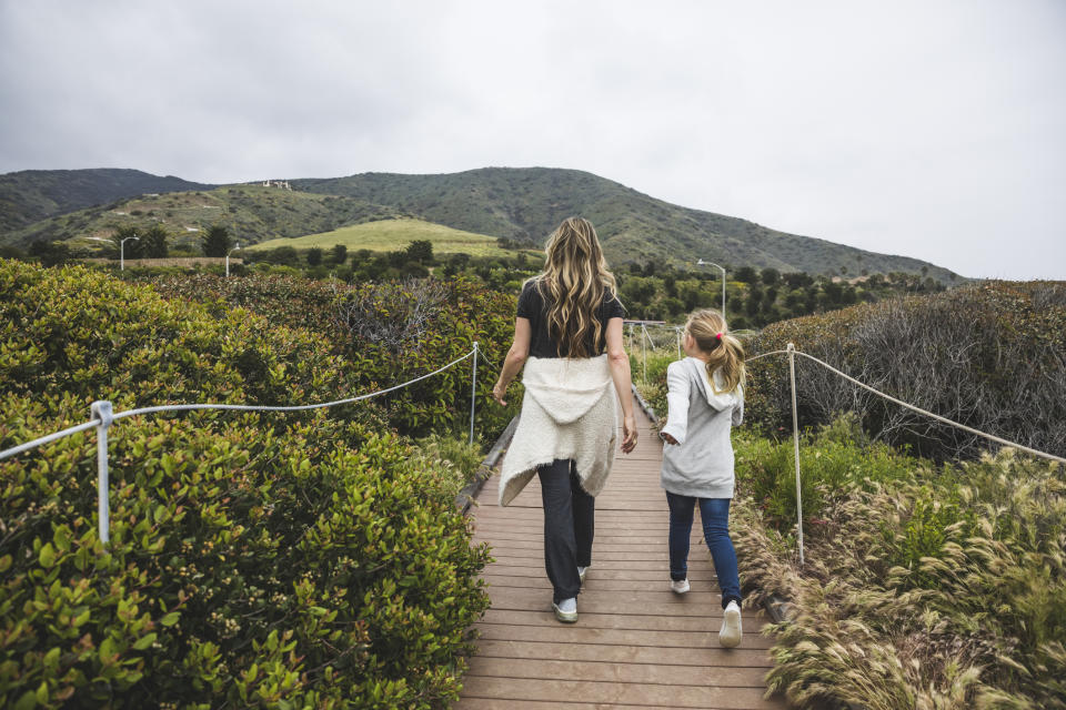 A woman and a child walk hand-in-hand along a wooden path through lush greenery, with mountains in the background