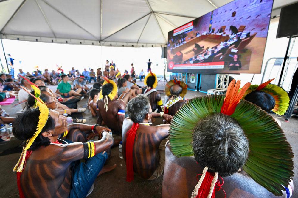 People wearing headdresses and displaying tattoos on their arms sit on the floor under a huge white marquee watching a large screen that shows events in a court.