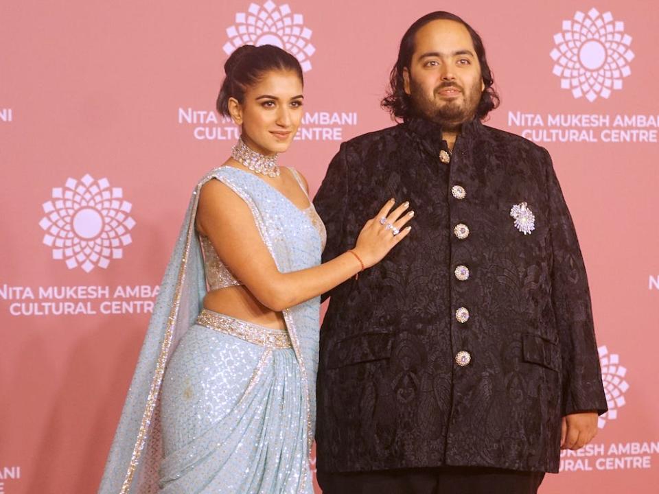 Anant Ambani wearing a black jacket and Radhika Merchant wearing a light blue sari