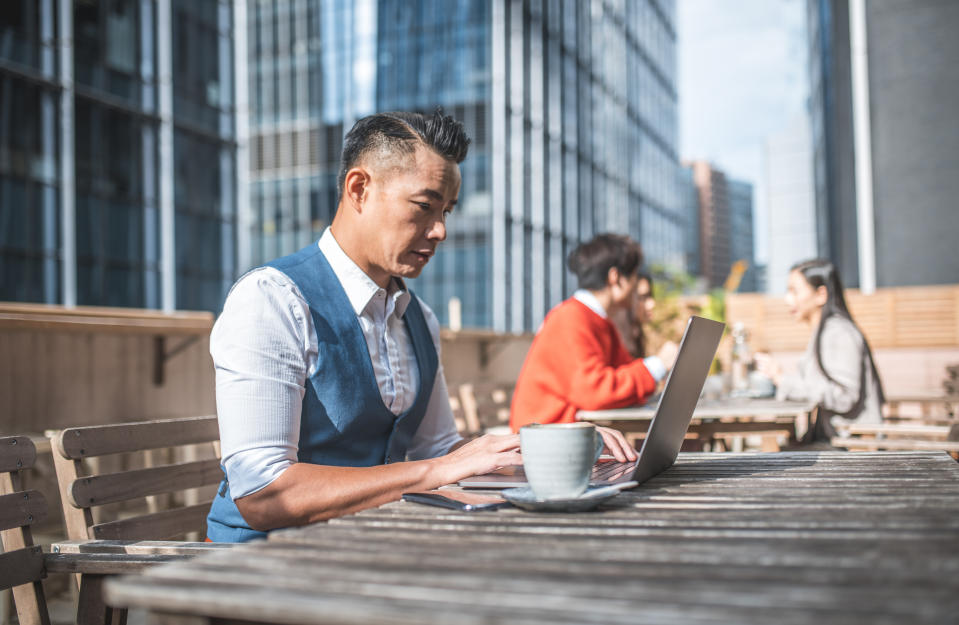 Side view of mature businessman drinking coffee while using technologies. Confident professional is sitting at table. He is working at outdoor cafe.