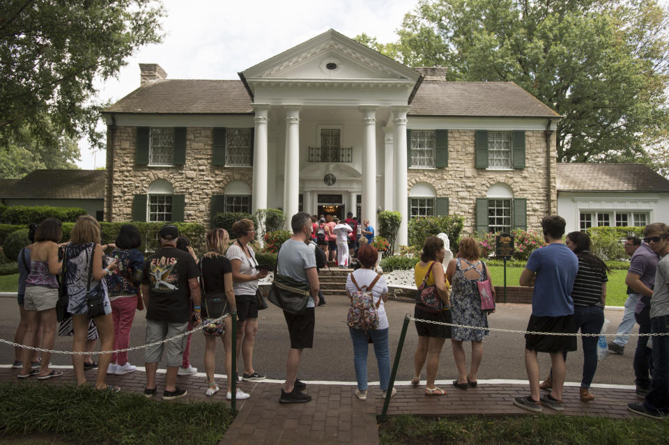 Several dozen people stand on the sidewalk across the street from a large stone house with four columns on either side of the entrance.