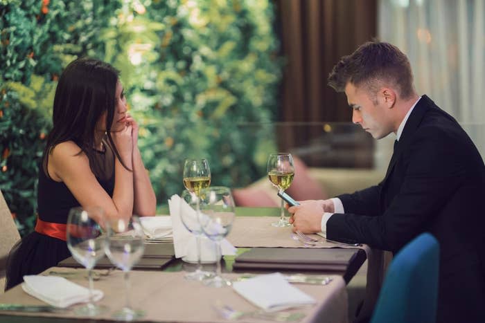 Two people at a dining table, a woman looking at a man who is focused on his phone, glasses of wine and dinnerware present