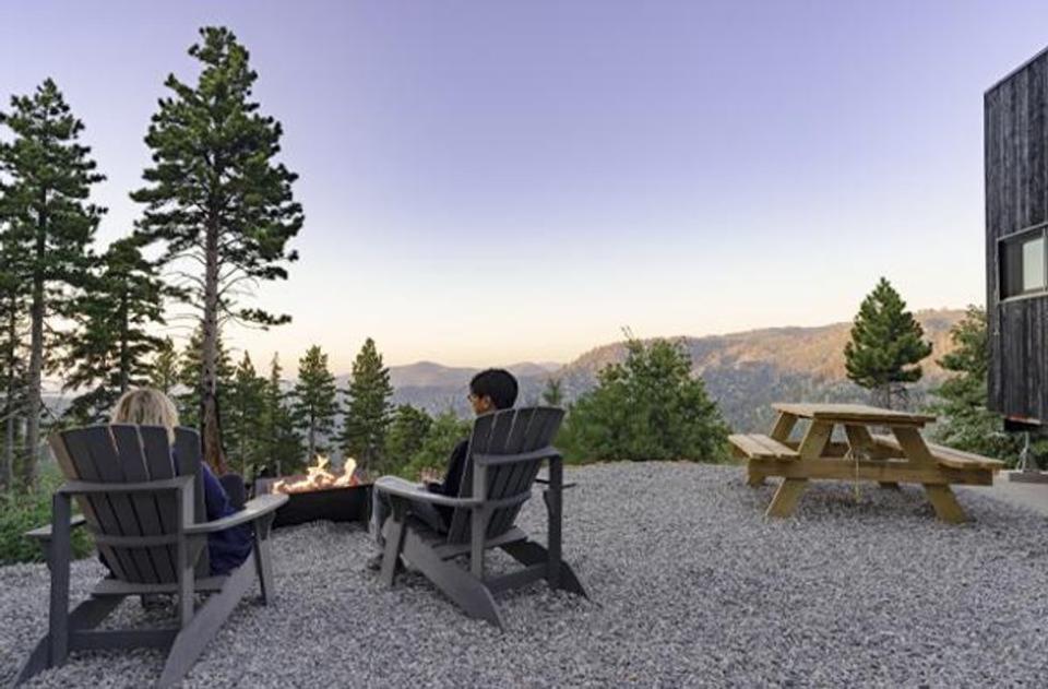 Two people sit next to a fire pit outside of their tiny house cabin