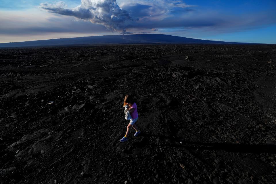 Abigail Dewar, of Alberta, Canada, holds a stuffed animal as she walks over hardened lava rock from a previous eruption as the Mauna Loa volcano erupts, behind, Wednesday, Nov. 30, 2022, near Hilo, Hawaii (AP)