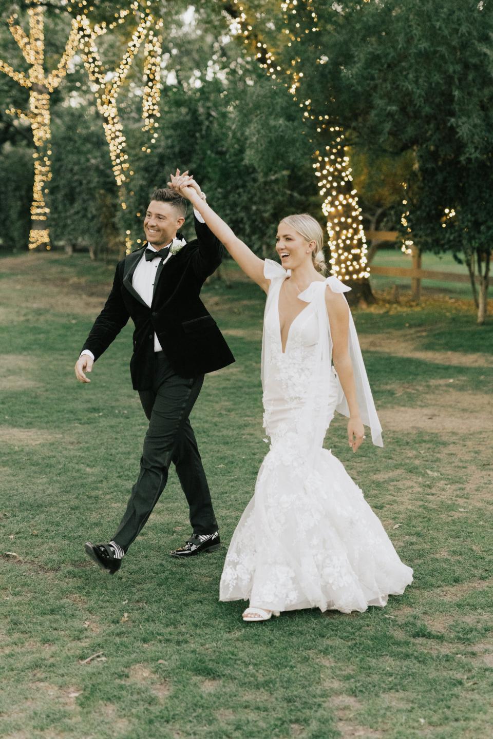 A bride and groom raise their hands and smile as they walk together.