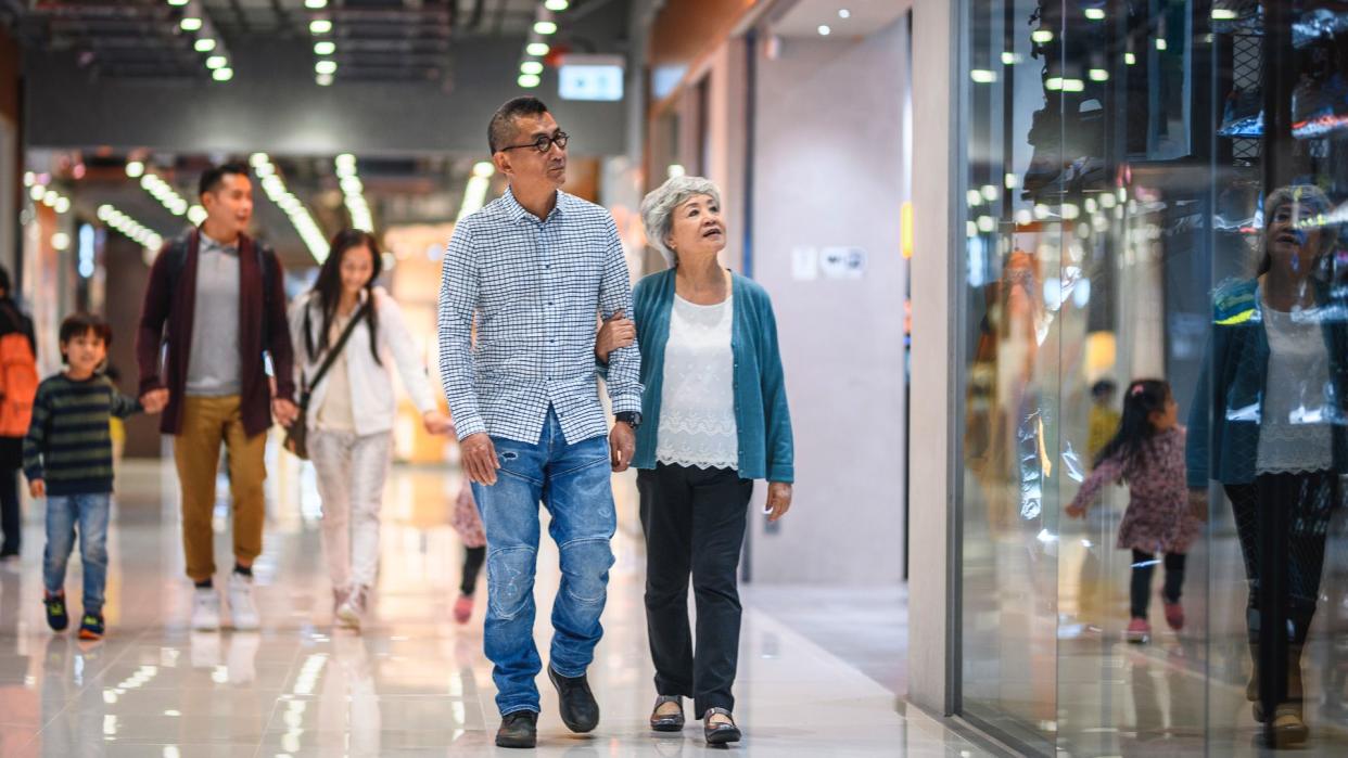 Senior Chinese man and woman walking arm in arm through Harbour City Shopping Centre with younger family members in background.