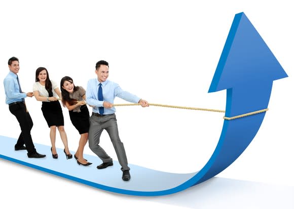 Four businesspeople tug on a rope to pull a large charting arrow upwards.