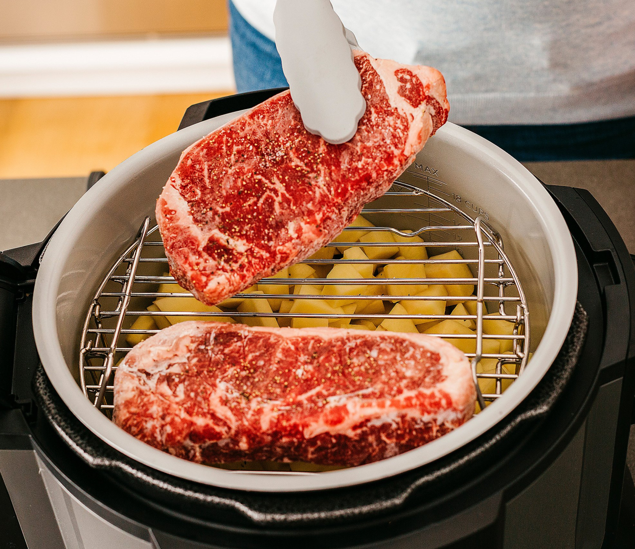 Person placing raw steak on grill of multi-cooker above cubed potatoes.