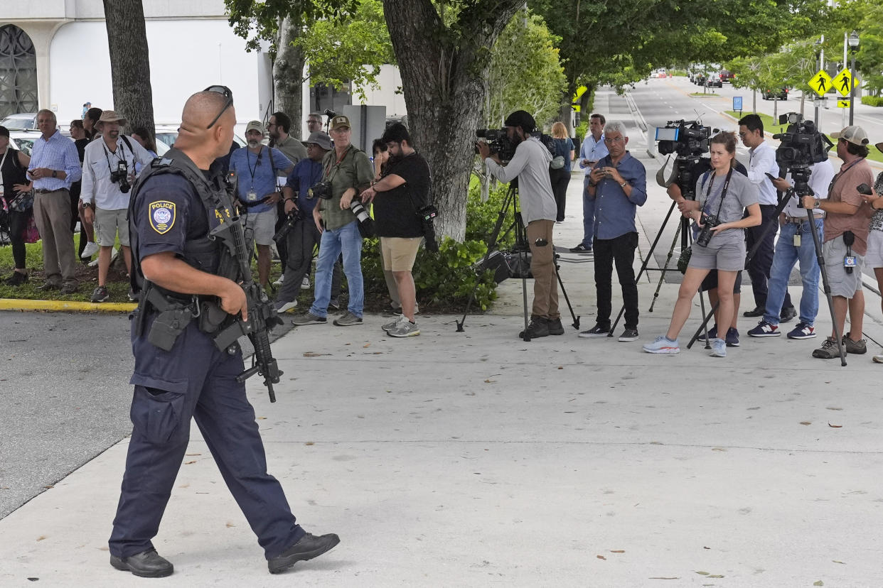 A Department of Homeland Security officer holding a weapon walks past a line of reporters and onlookers.