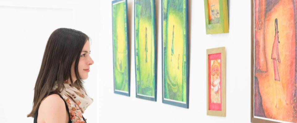 side view of young caucasian woman standing in an art gallery in front of colorful framed paintings displayed on a white wall