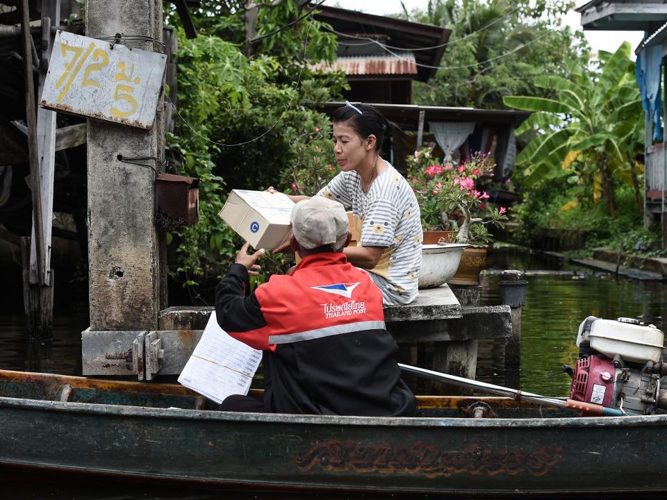 A man wearing a red uniform jacket sits in a boat withe a small motor and hands a package to a woman in Thailand.