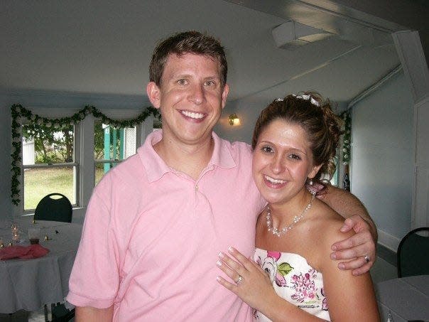 Terri Peters and her husband on her wedding day. Terri has her hair pulled back into a bun and wears a pearl necklace, pearl drop earrings, and a strapless white dress with pink roses on it. She has her hand on her husband's chest. Her husband stands to her right with his arm around her and wears a pink polo shirt. Behind them you can see they are standing in a room with tables that have white tablecloths and votive candles, along with a white rose garland along the window.