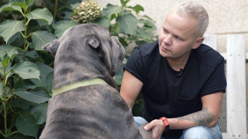 Francis sitting in black t-shirt, short blond hair, holding the paws of a large dog