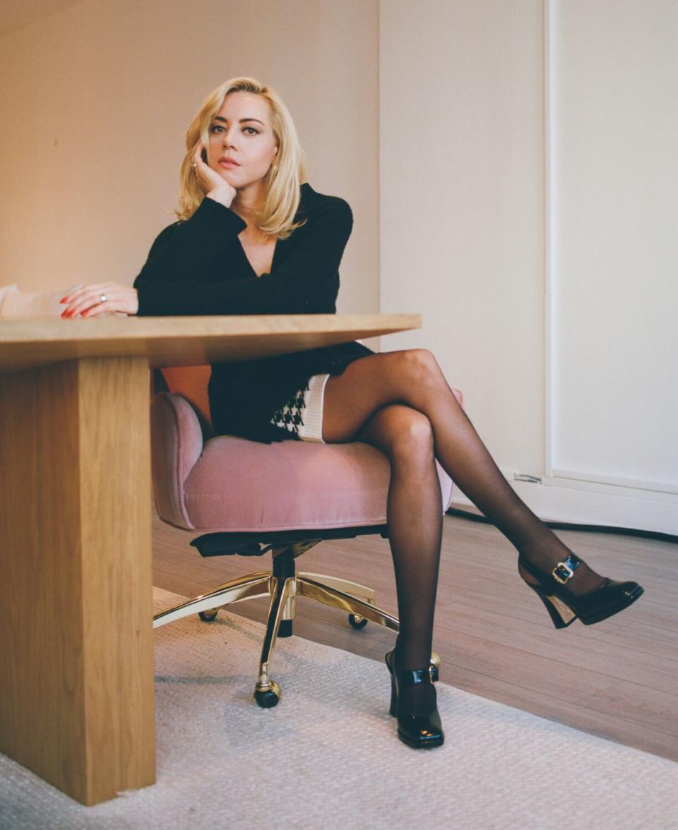 Aubrey Plaza sitting in a pink chair at a wooden table.