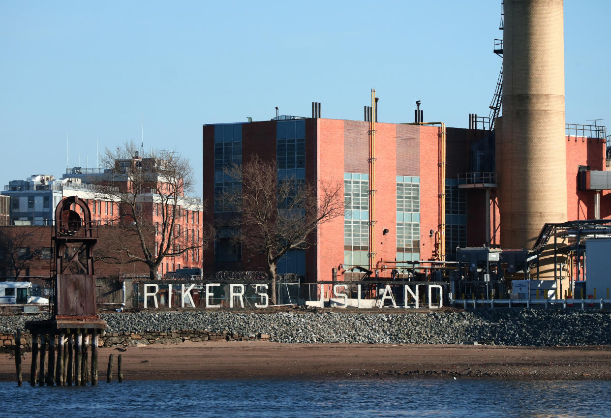 A sign says Rikers Island, with buildings of the Rikers Correctional Center in the background and the East River in the foreground, in New York City. 