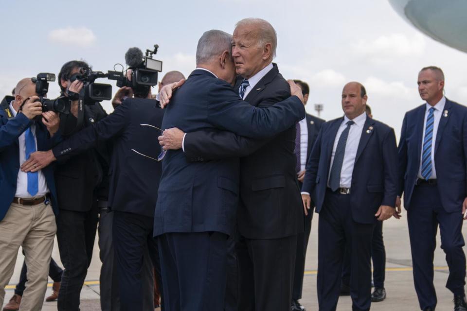 Two men in dark suits embrace, as other people stand around them, some holding cameras.