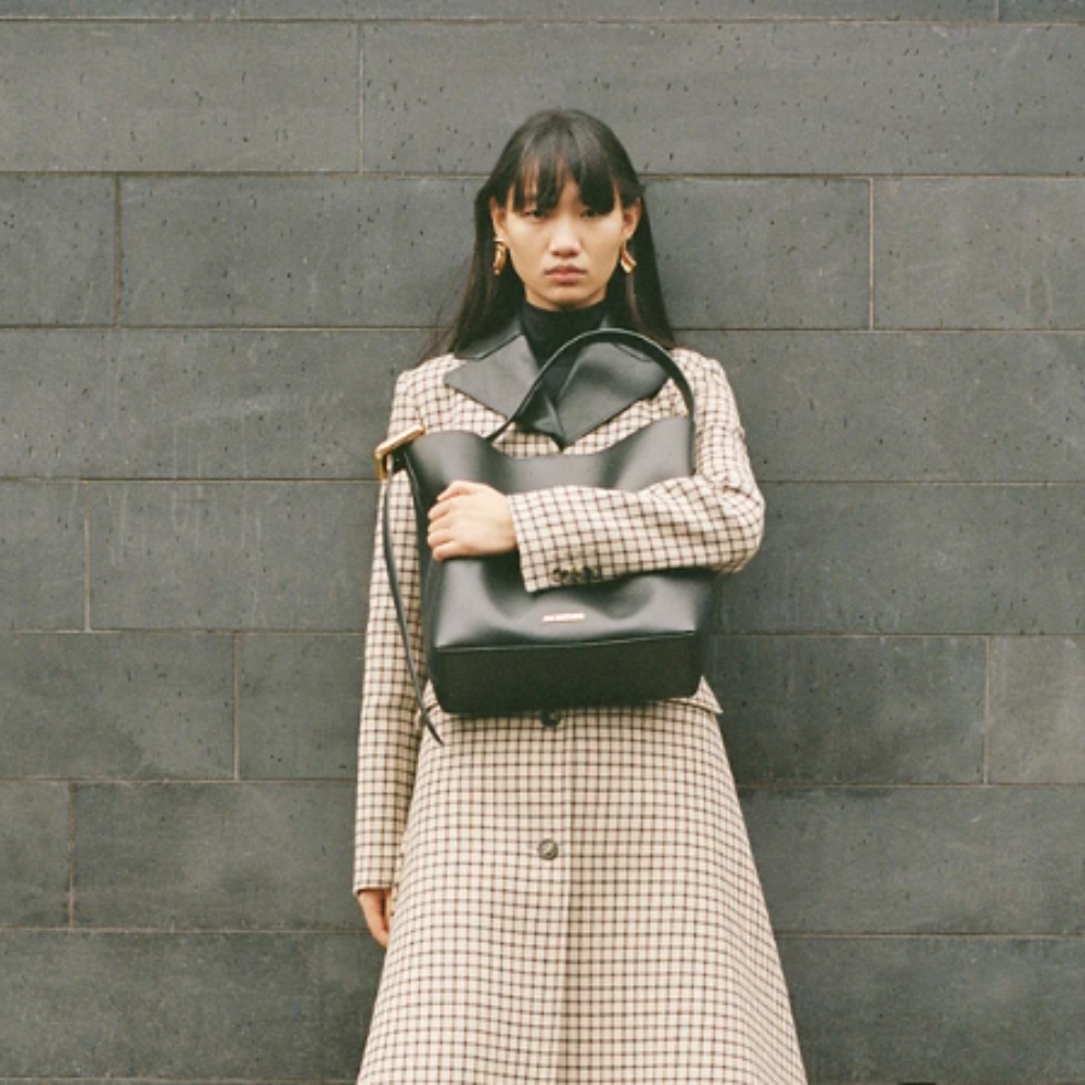  Woman in checked coat and gold earrings holding Selfridges bag with grey brick background. 