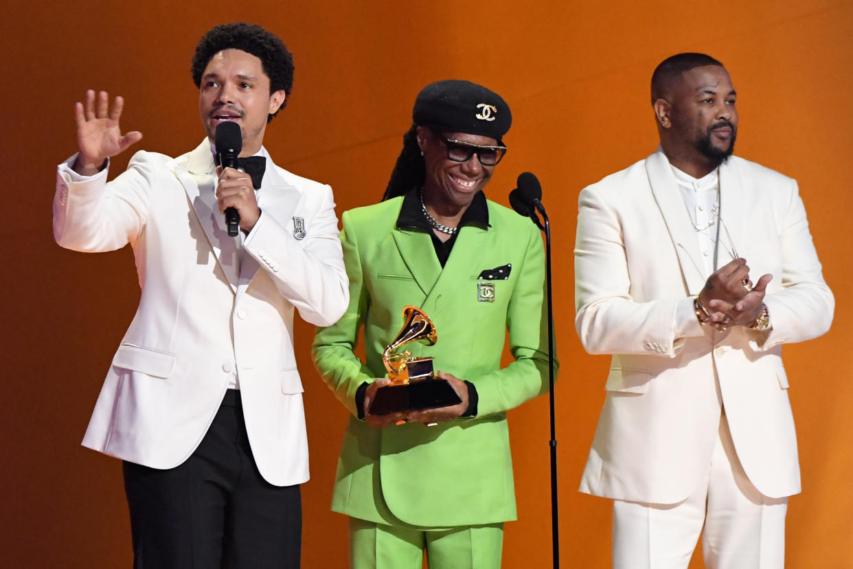 Trevor Noah speaks at the microphone, with Nile Rodgers holding Beyoncé's Grammy, and The Dream applauding.