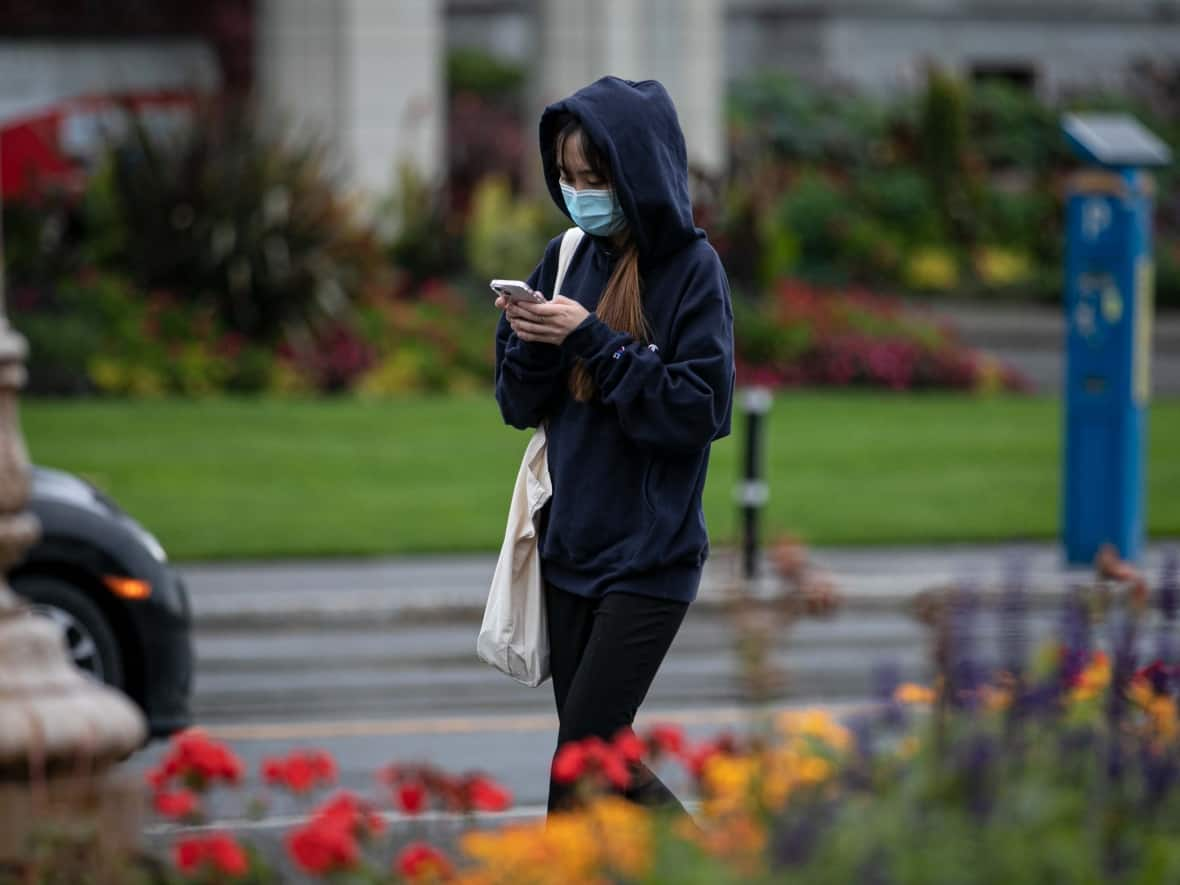 A pedestrian wearing a protective face mask looks at a cellphone while walking in Victoria, B.C. on Oct. 19, 2021. (Ken Mizokoshi/CBC - image credit)