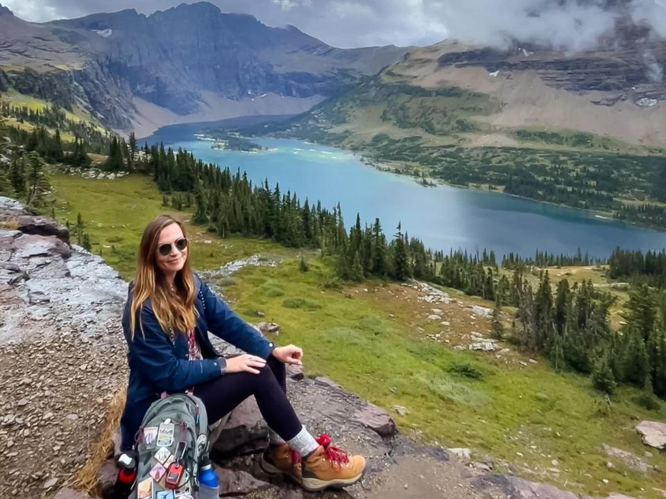 Emily sits on a rock, with her backpack covered in patches next to her. Behind her is a lake surrounded by mountains and trees.
