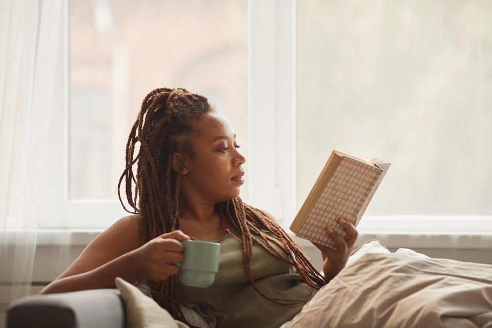 young woman lying on sofa drinking coffee and reading an interesting book at home