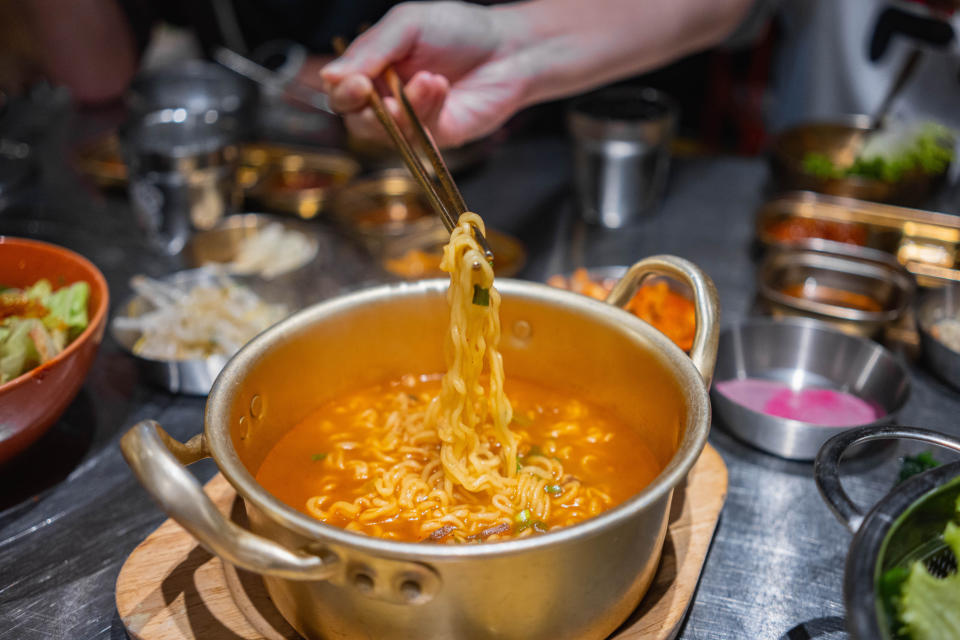 Person using chopsticks to lift noodles from a steaming pot of soup at a table with various dishes