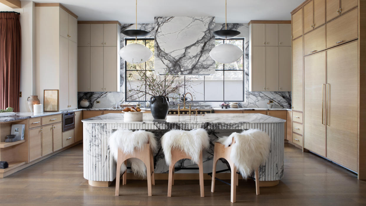  Kitchen with beige cabinetry, veined marble splashback and cooker hood, and round fluted marble island with wood bar stools 