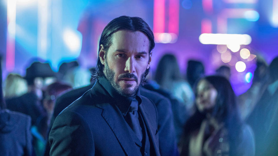  Keanu Reeves as John Wick, standing in a crowd of people with neon in the background in John Wick: Chapter 2 