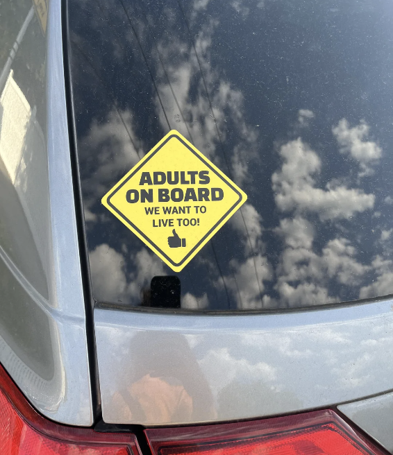 Sticker on a car window reads "ADULTS ON BOARD, WE WANT TO LIVE TOO!", parody of baby on board signs