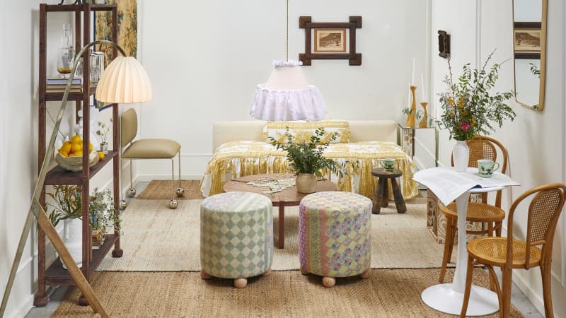 Head on view of a living room with white walls, a low couch with printed yellow and white ruffled cushions, two patchwork stools, and a small bistro table and chairs in the front right.