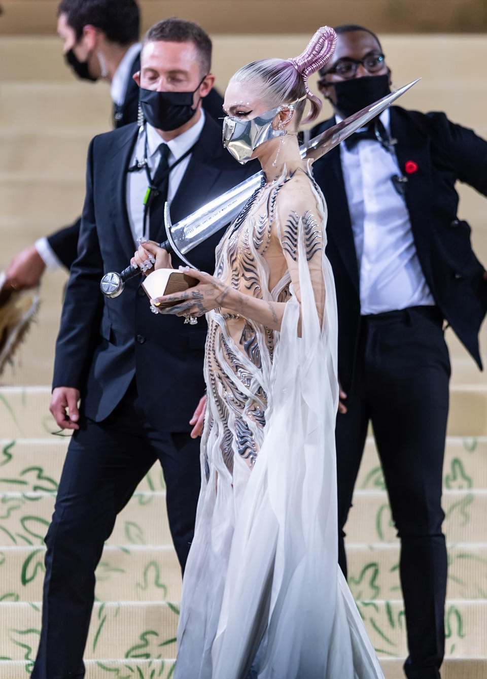 Grimes in a futuristic, sculptural white gown with metallic accents, at a formal event, with masked attendees in the background
