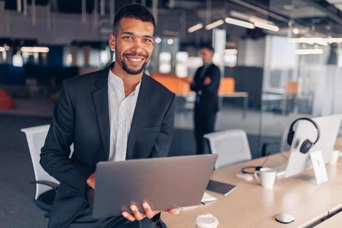 A smiling person in a business suit holding a laptop.