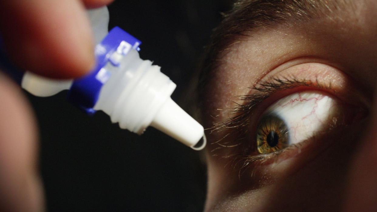  close up of a man's eye as he's squeezing a generic eye drop dropper near his face, pushing out a drop of clear liquid 