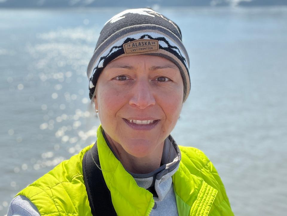 rebecca posing in front of a body of water and mountain scape wearing a winter hat and yellow vest