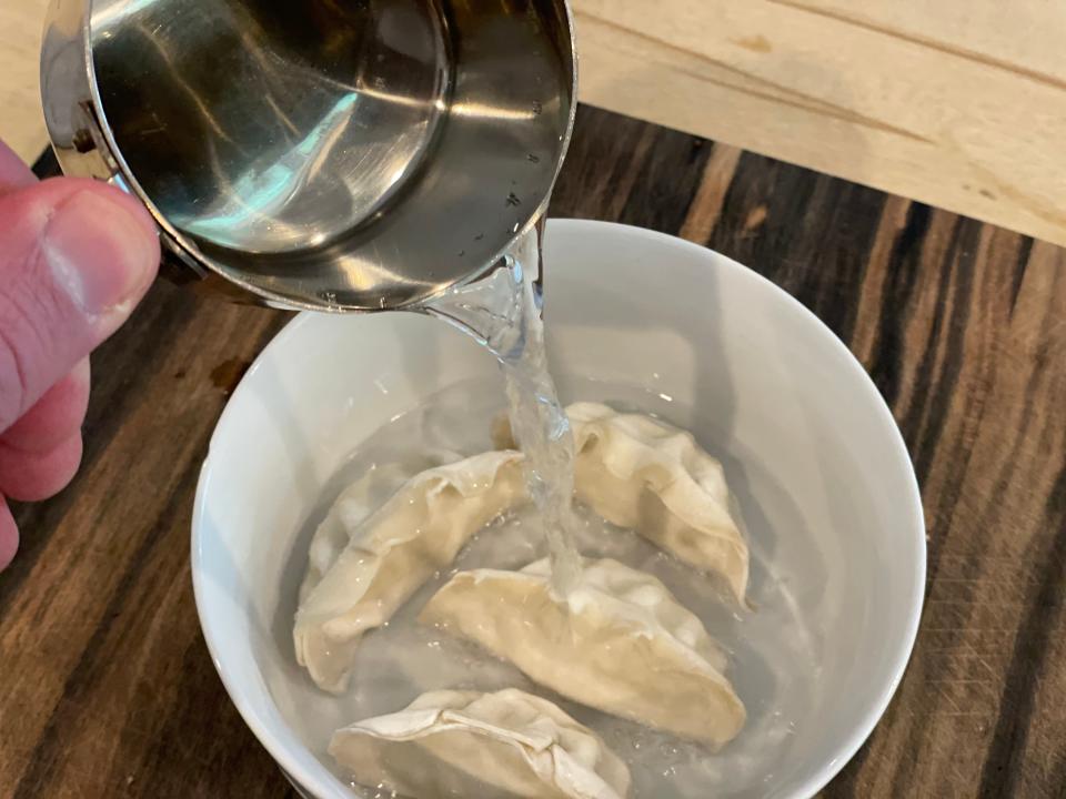 hand pouring a cup of water over a bowl with frozen potstickers in it