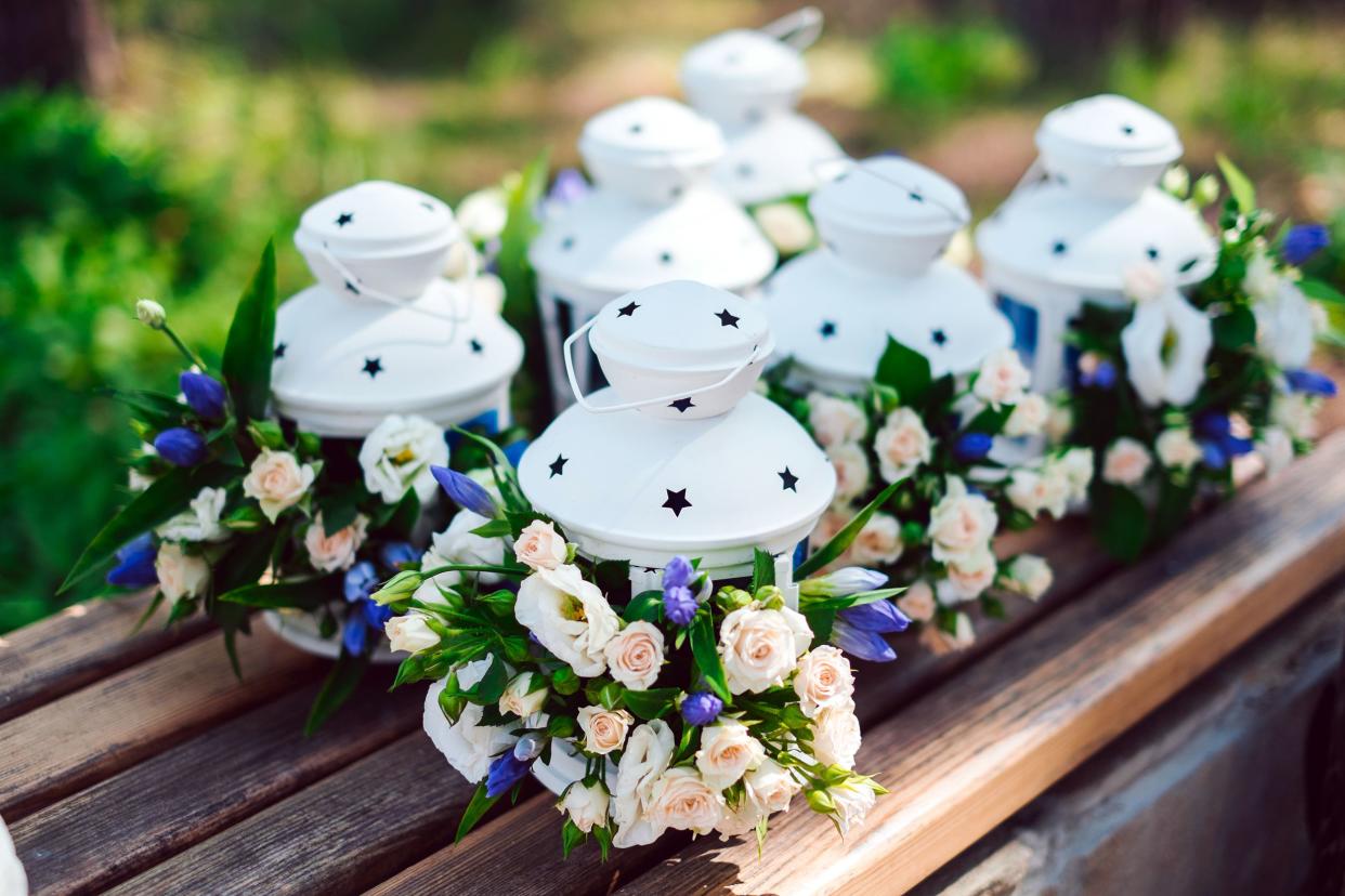 Six white lanterns decorated with small pink roses and blue flowers, selective focus, on a wooden plank, for wedding decorations, green plants blurred in the background