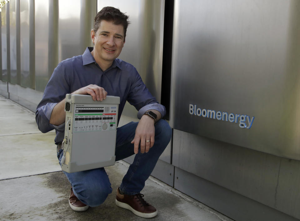 Joe Tavi, Bloom Energy senior director of manufacturing, holds a refurbished ventilator as he kneels beside fuel cells, Wednesday, April 1, 2020, in Sunnyvale, Calif. The new coronavirus outbreak has prompted companies large and small to rethink how they do business. Bloom Energy, in San Jose, Calif., makes hydrogen fuel cells. But recently, they have been refurbishing old ventilators so hospitals can use them to keep coronavirus patients alive. (AP Photo/Ben Margot)