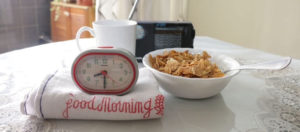 Grey and red coloured alarm clock showing 8.30, on top of a "Good Morning" towel, beside a white bowl of cereal, a white mug of coffee and a black radio.