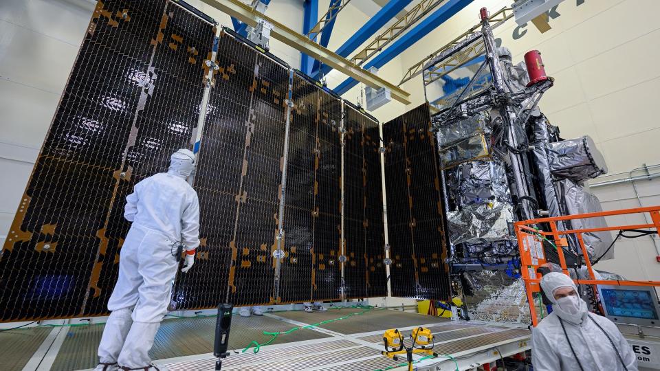 engineers in white clean suits look up at a large solar panel extending from a silver cube-shaped spacecraft in a hangar