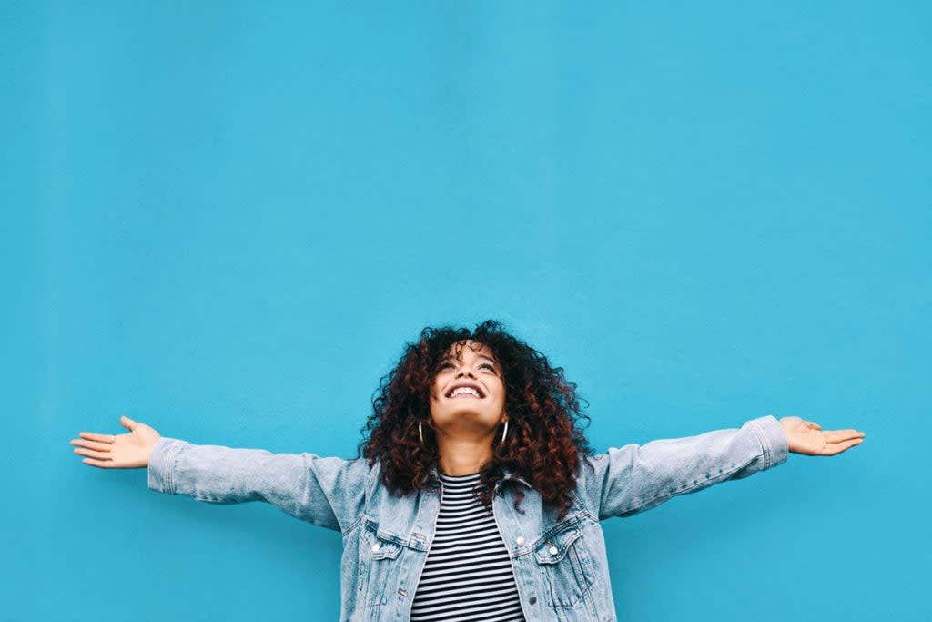 A woman throws her hands in the air and smiles upward against a blue wall outside.