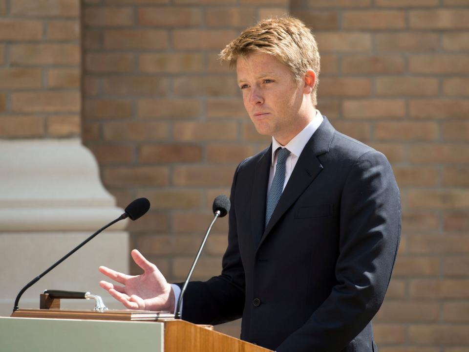 Hugh Grosvenor standing behind a podium and speaking while wearing a black suit and blue tie.