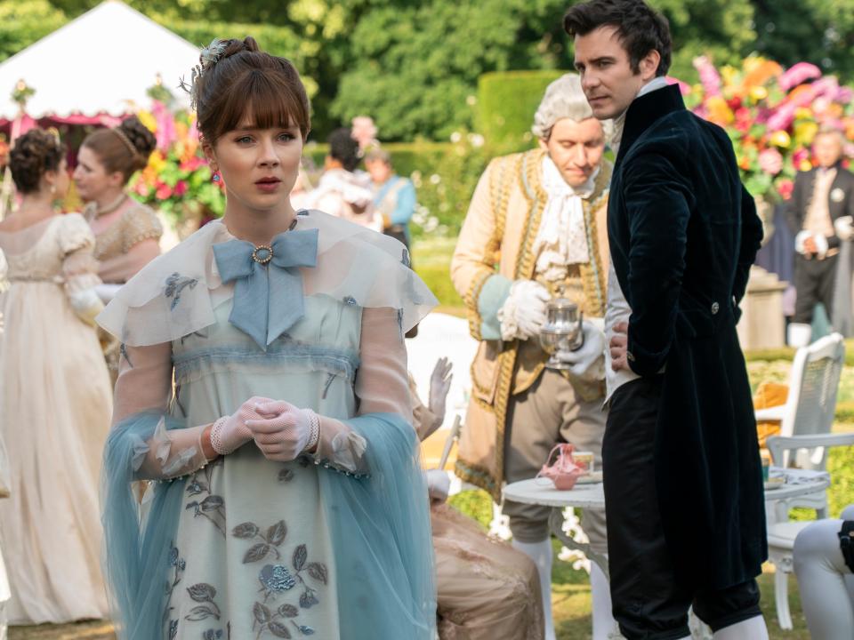 eloise bridgerton in a blue gown with a bown at her neck, looking bemused while standing at a garden party