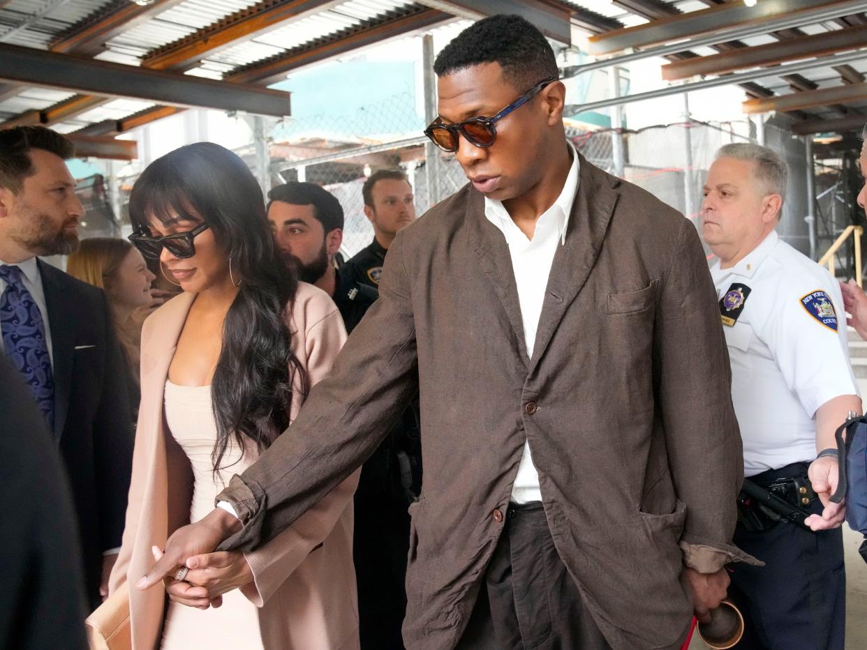 Jonathan Majors leaving a courthouse while wearing a brown jacket and a white shirt and holding the hand of a woman who is wearing a light pink dress.