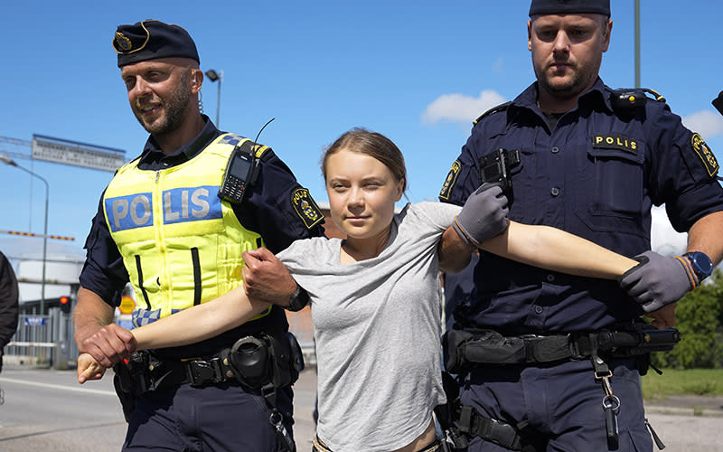 Climate activist Greta Thunberg is detained by police during a protest. There are two guards, one on either side of her, securing her arms straight out to the side by securing one hand under the armpit and holding her wrist with the other.