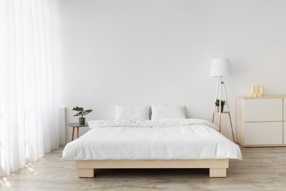 Minimalist bedroom with a low wooden bed frame, white bedding, a bedside table with plants, a floor lamp, and a dresser with candles and decor items