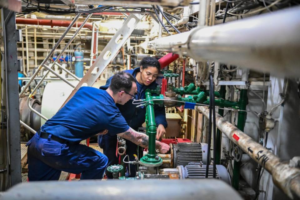 Two Coast Guard crew members, a man and a woman, work amid pipes on the ship.