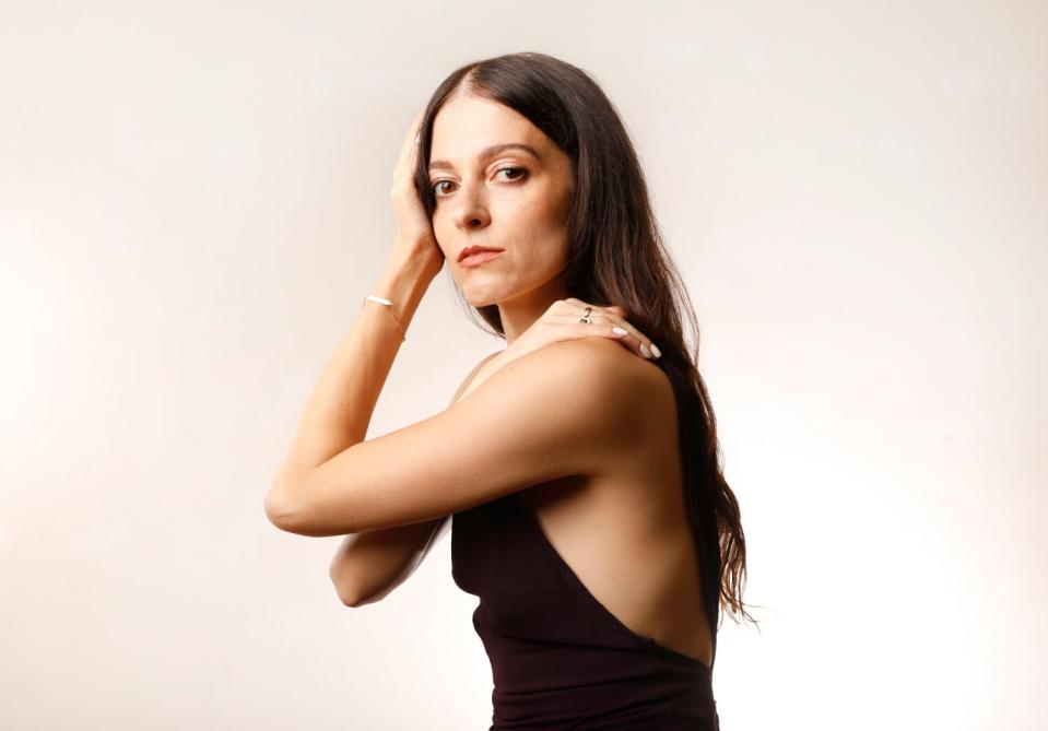 A woman with long brown hair, her arms crossed with her hands on her head and shoulder.