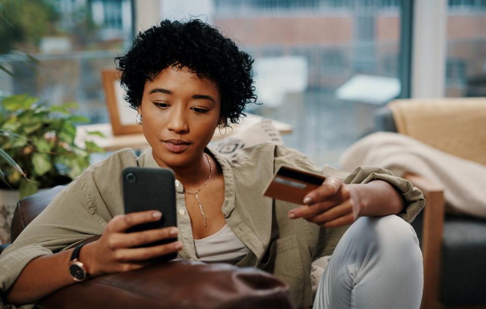 Shot of a young woman holding her credit card while using her cellphone at home.