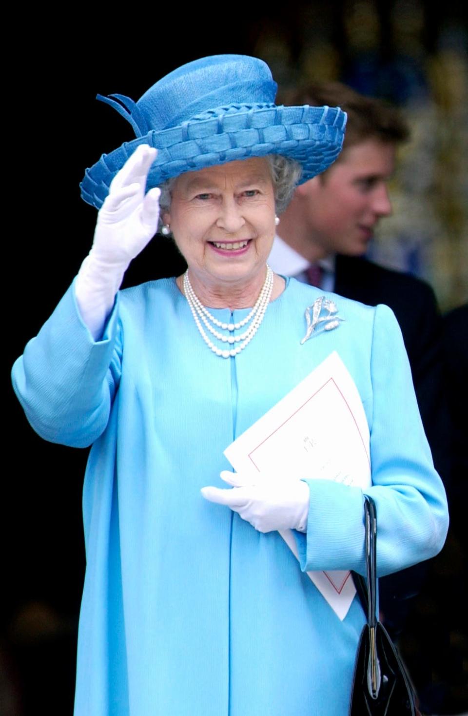 The Queen smiling and waving in a bright teal blue long sleeve overcoat with a woven wide-brimmed matching hat. She has on pearl earrings and necklaces and white gloves.