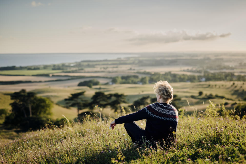 A person is sitting on a grassy hillside, overlooking a vast, scenic landscape with fields and a distant body of water